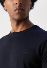 Navy sweater with a crew neckline and ribbed trim, featuring a subtle embroidered logo on the chest. Fabric has a smooth texture.