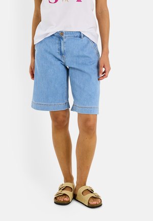 Persona que lleva puestos unos pantalones cortos bermuda vaqueros azul claro, una camiseta blanca y sandalias beige sin cierre, de pie frente a un fondo blanco liso.