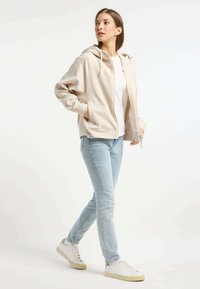 Beige geripptes Zip-Hoodie mit Kapuze, getragen über einem weißen Shirt, kombiniert mit hellblauen Jeans und weißen Sneakers. Glatte Stoffoberfläche.