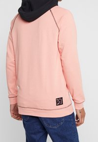 Felpa rosa con cappuccio nero, dotata di maniche lunghe, cuciture a contrasto e un'etichetta con logo sul lato inferiore. Tessuto morbido.