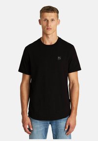 Schwarzes Baumwoll-T-Shirt mit Rundhalsausschnitt, kurzen Ärmeln und einem kleinen Logo-Patch auf der linken Brust. Getragen mit hellblauen Jeans.