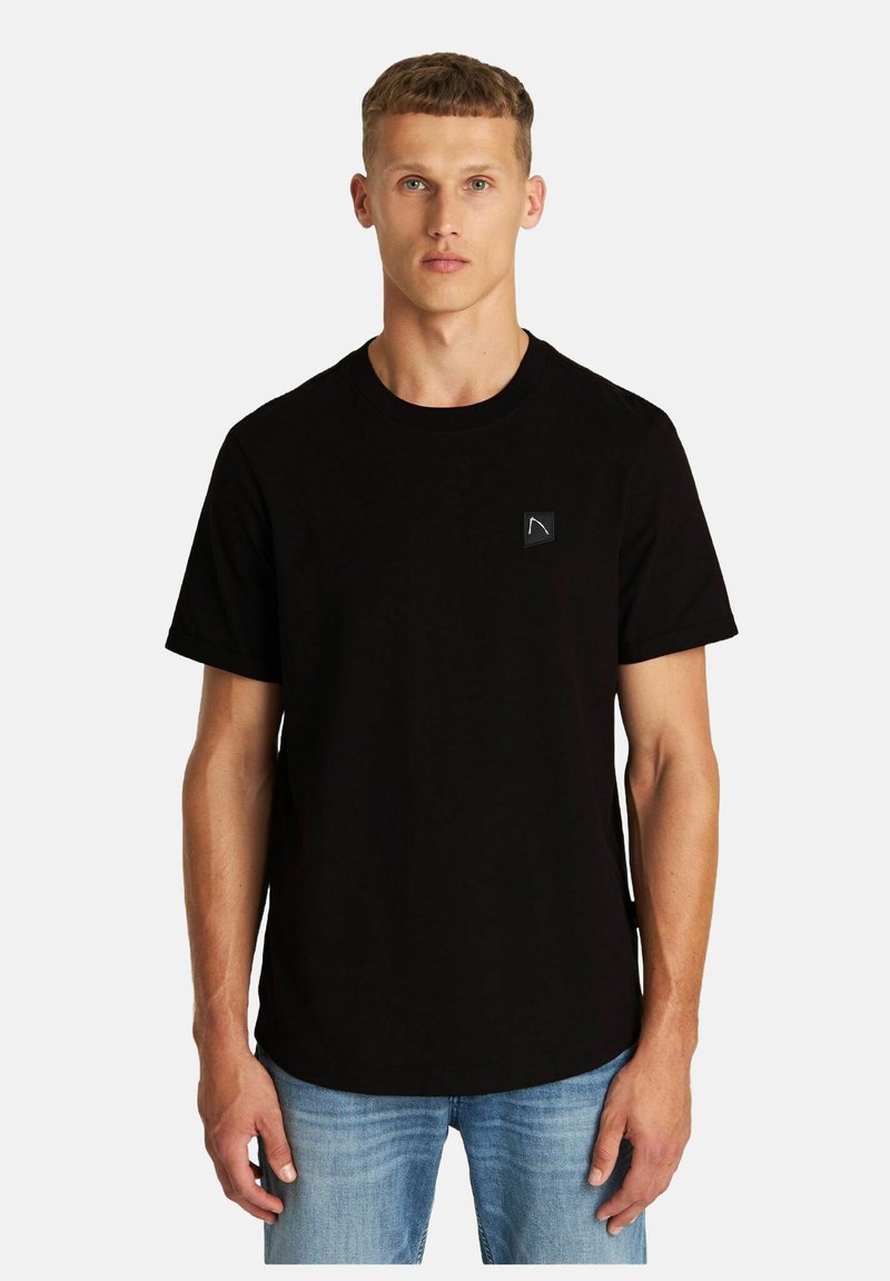 Schwarzes Baumwoll-T-Shirt mit Rundhalsausschnitt, kurzen Ärmeln und einem kleinen Logo-Patch auf der linken Brust. Getragen mit hellblauen Jeans.