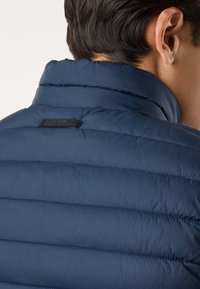 Uomo con capelli corti e scuri che indossa una giacca trapuntata blu navy visto da dietro, con particolare attenzione al colletto e alla parte superiore della schiena.