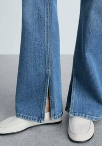 Vaqueros de denim azul de pierna ancha con aberturas laterales en el dobladillo, que presentan detalles de costura ligera. Combinados con zapatos planos de suela clara.