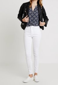 Giubbotto in pelle nera da motociclista, blusa a fantasia blu navy, jeans skinny bianchi e scarpe slip-on bianche, che mostrano un outfit casual e stratificato.