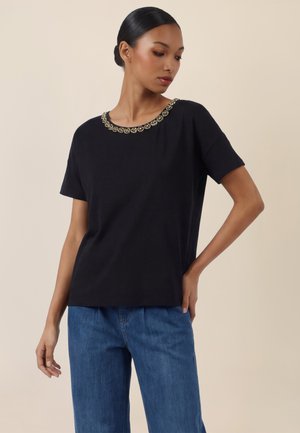 Woman wearing black short-sleeve top with beaded neckline and blue jeans, posing with one hand on hip against plain background.