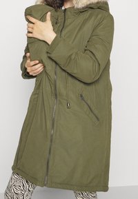 Parka verde oliva con capucha forrada de piel sintética, cierre frontal con cremallera y bolsillos laterales. El material parece resistente con una textura suave.