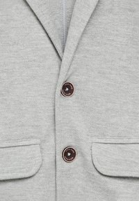 Ljusgrå barnblazer med två bruna knappar märkta "MNG KIDS" och två framfickor med klaff.