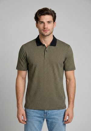 Giovane uomo in piedi, rivolto verso l'obiettivo, che indossa una polo a maniche corte verde oliva con motivo, colletto nero e jeans azzurri.