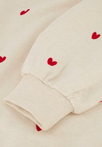 Cremfarbener Pullover aus Stoff mit rot gestickten Herzen, ausgestattet mit gerippten Bündchen und weicher Textur, die ein entspanntes Design für optimalen Komfort zeigt.