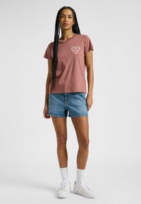Camiseta de algodón color rosa con mangas cortas, que presenta un gráfico de un corazón. Combinada con pantalones cortos de mezclilla azul claro y zapatillas blancas.