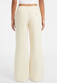 Donna che indossa pantaloni crema a vita alta e gamba larga con tasche posteriori e un piccolo logo sulla tasca destra, in piedi di spalle.