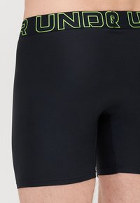Schwarze Boxershorts aus elastischem Material mit breitem, elastischem Bund, der auffälligen grünen Text trägt. Glatte Textur mit passgenauer Form.