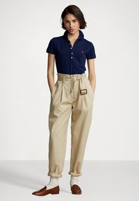 Μπλε πουκάμισο πόλο με γιακά και πατιλέτα με κουμπιά, σε συνδυασμό με μπεζ παντελόνια ψηλόμεσα με ζώνη και γυρισμένα μανίκια. Καφέ πλεκτά μοκασίνια και λευκές κάλτσες.