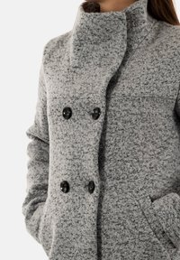 Manteau en mélange de laine gris avec un col haut, deux boutons noirs et des poches latérales ; tissu texturé avec un motif moucheté.