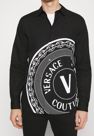 Homme portant une chemise noire boutonnée avec un grand logo rond blanc Versace Couture et un motif chaîne à l'avant.