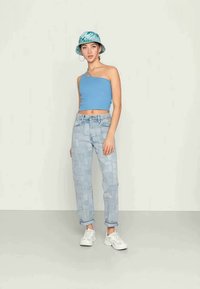 Top cropped azul claro de um ombro só emparelhado com jeans de lavagem clara com remendos, complementado por um chapéu de balde verde com padrão e ténis brancos.