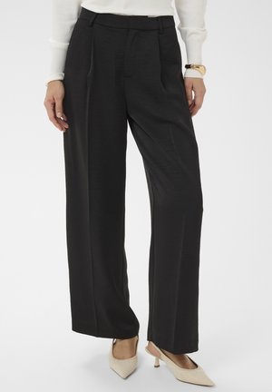 Žena v čiernych širokých nohaviciach s vysokým pásom, bielych lodičkách so špicatou špičkou a bielom top s dlhým rukávom a zlatým náramkom.