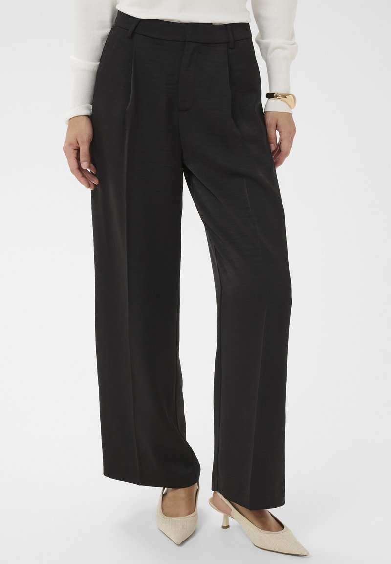 Femme portant un pantalon noir taille haute à jambes larges, des talons blancs à bout pointu et un haut blanc à manches longues avec un bracelet en or.