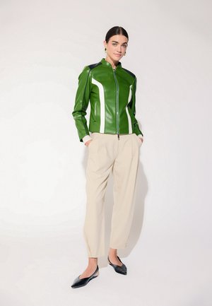 Mujer con cabello oscuro con una chaqueta de cuero verde con rayas blancas, pantalones beige, zapatos planos negros puntiagudos y pendientes verdes, de pie con las manos en los bolsillos.
