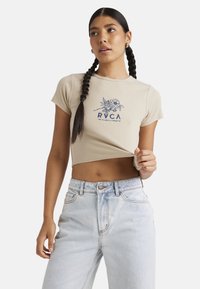 Beige cropped t-shirt with short sleeves, featuring a blue floral graphic and logo design. Worn with light-wash high-waisted jeans.