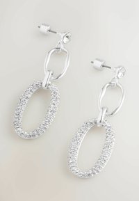 Stříbrné visací náušnice se spojenými oválnými tvary, jeden hladký a druhý osázený malými průhlednými krystaly, vystavené na světlém pozadí.