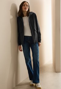 Lätt, svart jacka med huva och fickor, tillsammans med en vit t-shirt och blå utsvängda jeans. Modellen bär vita sneakers.