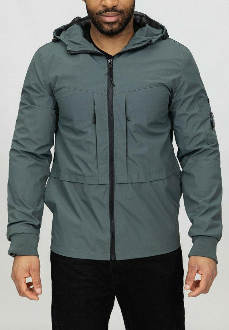 Veste verte zippée avec une capuche, dotée de deux poches avant et de poignets côtelés. Tissu lisse avec une texture subtile et des accents noirs.