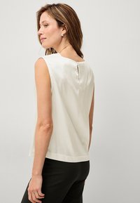 Ärmellose weiße Bluse mit rundem Halsausschnitt, ausgestattet mit einem kleinen Schlitz auf der Rückseite. Glatte Textur und lockere Passform, kombiniert mit dunklen Hosen.
