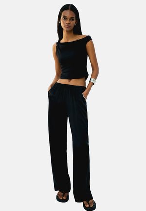 Top noir court avec un design dénudé des épaules, associé à un pantalon noir ample. Texture de tissu lisse, style minimaliste et détails discrets.
