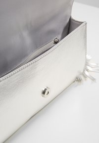 Sølvmetallisk clutch-taske med en glat tekstur, der har en front snap-lukning og en indvendig lynlåslomme. Accenter med frynsede kanter.