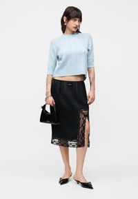 Femme portant un pull en maille torsadée bleu clair court, une jupe midi en satin noir avec une fente en dentelle, un sac à main noir et des mules à talons pointues noires.