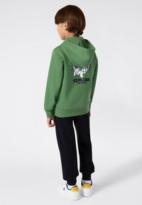 Felpa verde con una grande grafica bianca di un astronauta sulla schiena, pantaloni neri e scarpe bianche con dettagli gialli. Design morbido e casual.