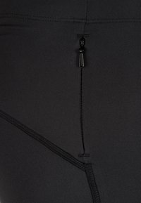 Detailní záběr na černou látku, na které je malý černý zip s viditelným šitím na hladkém a elastickém materiálu.