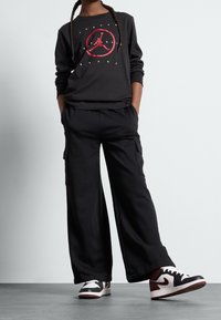 Czarna koszula z długim rękawem z czerwonym logo graficznym, w zestawieniu z czarnymi spodniami cargo z bocznymi kieszeniami oraz czarno-białymi trampkami.