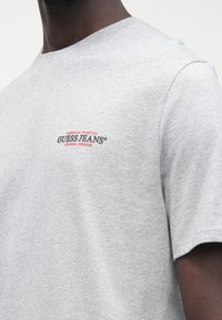 Szürke rövid ujjú pamut póló, kerek nyakkivágással, a bal mellrészen piros-fekete "GUESS JEANS" logóval.