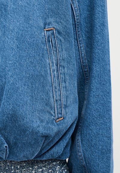 Gros plan sur une poche latérale d'une veste en denim bleu avec des coutures beiges et une ceinture élastiquée, sur un fond en tissu texturé.