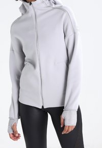 Chaqueta de punto gris claro con capucha, con detalles de mangas acanaladas y costuras planas, combinada con leggings negros ajustados.