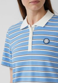 Eine Frau trägt ein blau-weiß-schwarzgestreiftes Poloshirt mit einem weißen Kragen und einem runden Aufnäher mit der Aufschrift "Tu, was du liebst" auf der Brust.