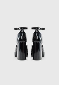 Sapatos de salto alto em couro verniz preto com um salto bloco robusto, detalhe de tira para o tornozelo e uma textura lisa e brilhante. Design de bico arredondado reconhecível.