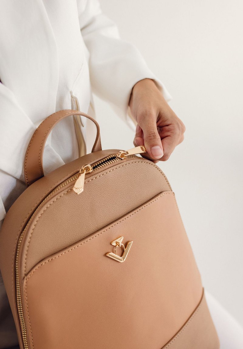 Hnědý kožený batoh se zlatým zipem a kováním. Zaoblený tvar s přední kapsou. Minimalistická akcentace loga na přední části.