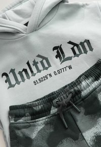 Szary hoodie z grafiką "Unltd Ldn" oraz współrzędnymi, w połączeniu z ciemnymi dresami w kamuflażu, z elastycznym pasem i sznurkiem.