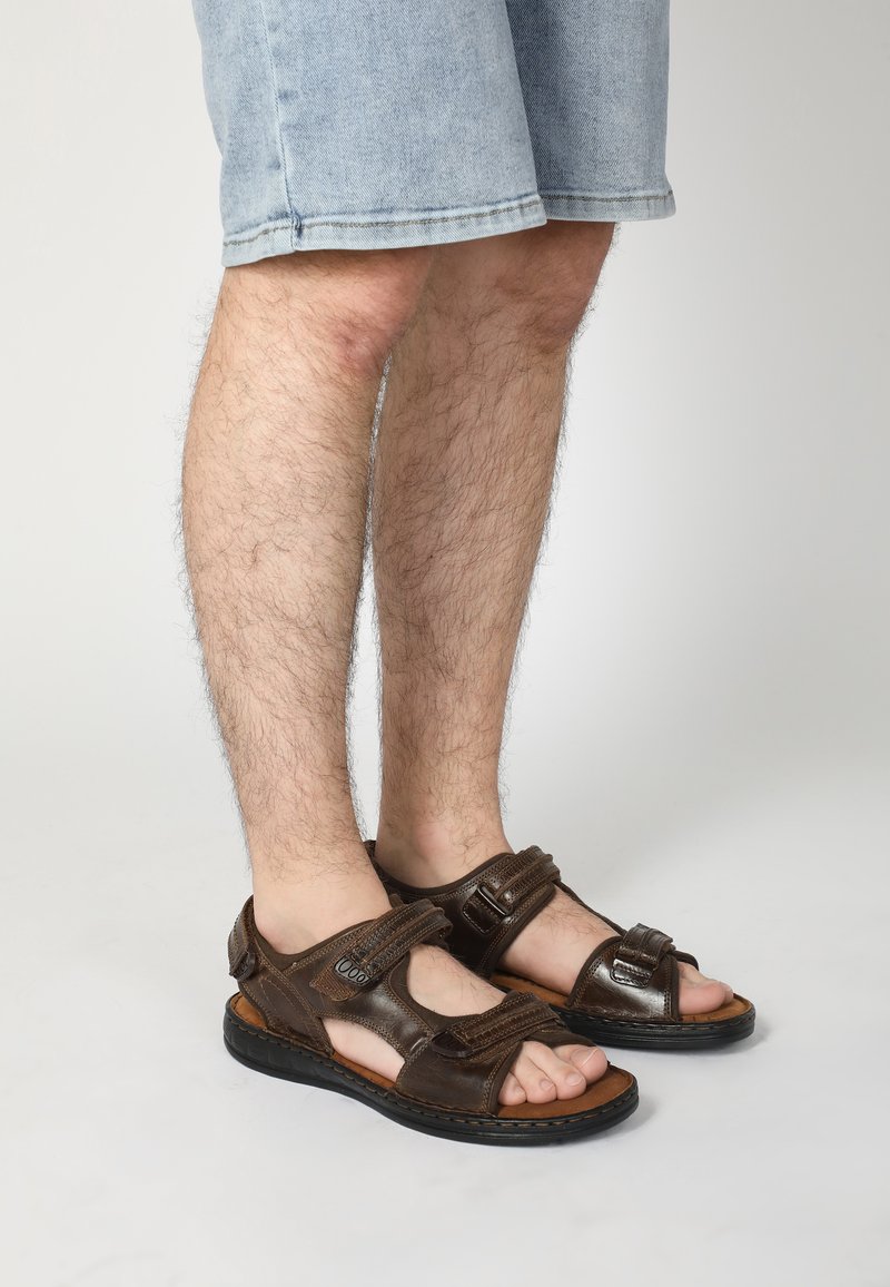 Braune Ledersandalen mit verstellbaren Riemen, die eine flache Sohle und ein offenes Zehendesign aufweisen, getragen mit hellblauen Jeansshorts.