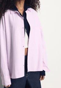 Camicia button-up a righe rosa chiaro con vestibilità morbida, dotata di tasca sul petto e maniche lunghe, indossata sopra un hoodie blu navy.