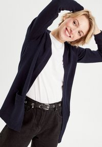 Cardigan azul marino con un diseño de punto texturizado, de frente abierto y con dos bolsillos, llevado sobre una camiseta blanca simple y pantalones negros.