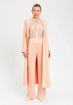 Donna in piedi che indossa un cappotto lungo color pesca, pantaloni a gamba larga e blusa di pizzo con fiocco, in posa su sfondo bianco.