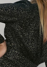 Blouse noire avec un motif animal subtil de couleur olive foncé. Elle présente des manches longues, une coupe ample et un tissu léger et texturé.