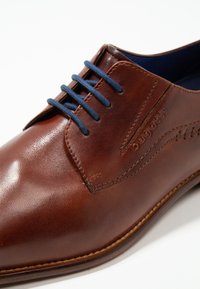 Chaussure en cuir marron avec une finition brillante, dotée de lacets bleus, de coutures latérales et d'un logo de marque en relief. Design à bout rond.
