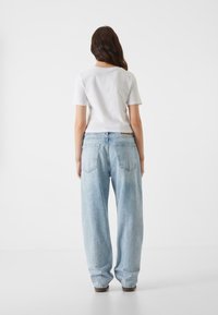 Camiseta blanca de manga corta y pantalones vaqueros acampanados de tiro alto en azul claro, con bolsillos traseros, vistos desde la parte de atrás sobre un fondo liso.