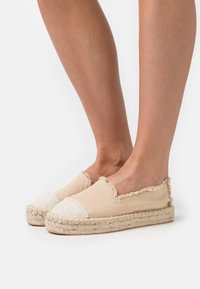 Pieds portant des espadrilles en toile beige avec des bords effilochés et des semelles en jute tressé sur fond blanc.